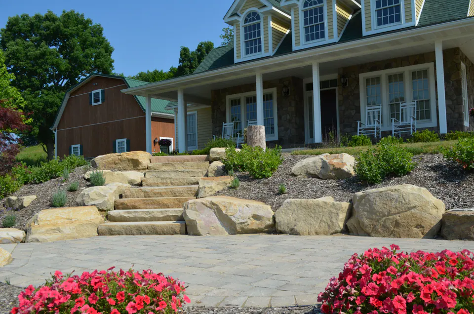 Broad stone steps between large boulders leading up to a stone and wood house with red annuals below